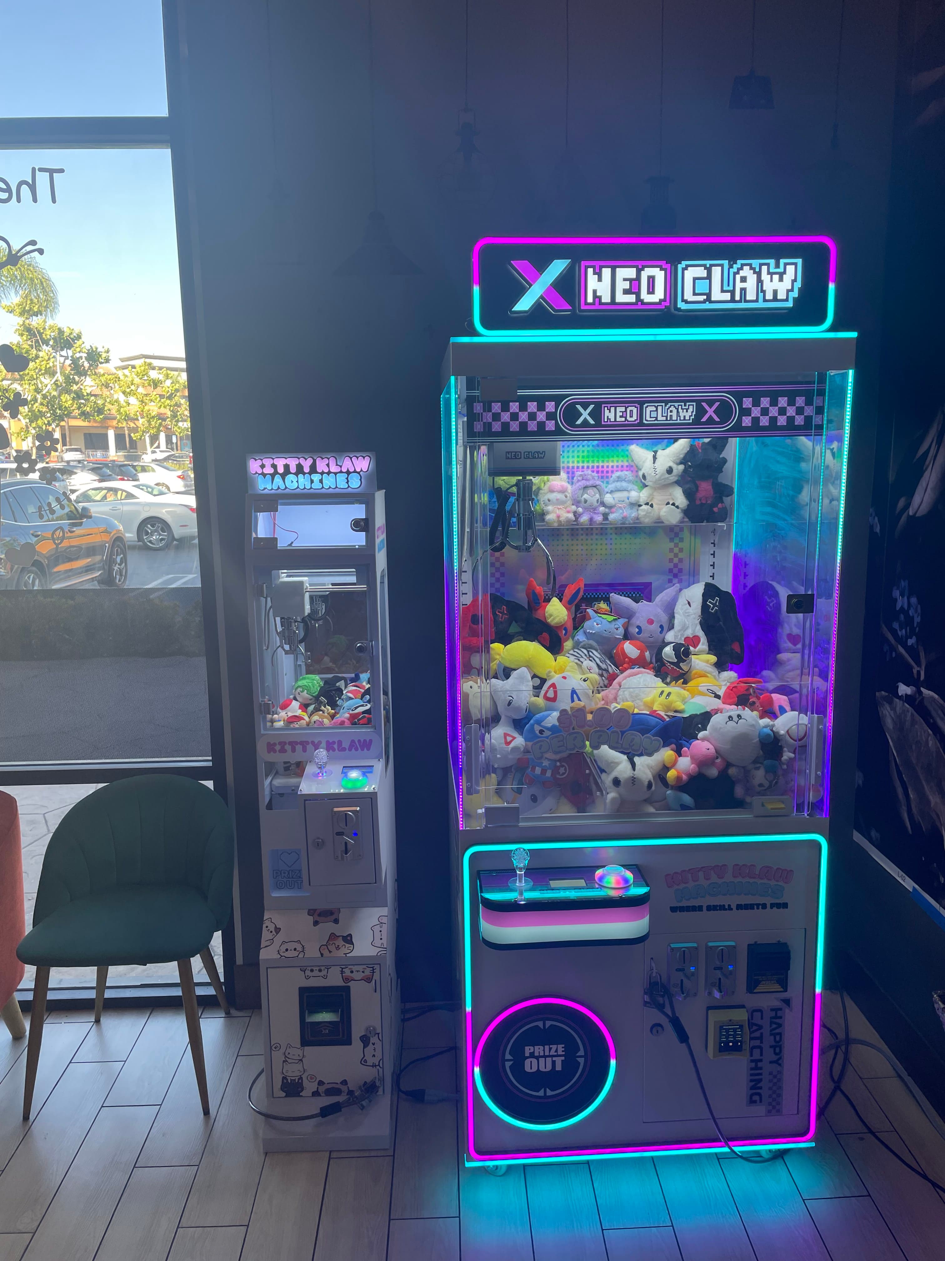Two neon-lit claw machines filled with plush toys stand beside a green velvet chair.