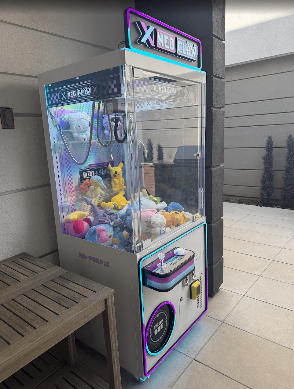 White Neo Claw machine with neon accents filled with plush toys on a tiled outdoor patio.